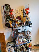 Full view of metal rooster sculptures on the top shelf with smaller ceramic and glass figurines placed on lower shelves along with framed rooster prints.