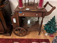 Front angled view of dark wooden tea cart showing drawer with white knobs, large wooden spoked wheels, lower shelf with glass cake stand, and side storage compartment with brass railing.
