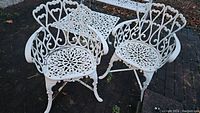 Two white wrought iron patio chairs on brick patio, showing decorative backs and seats, curved arms, and splayed legs.