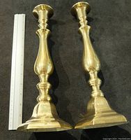 Two tall bronze/brass candlestick holders side by side next to a white ruler showing their size, hexagonal base bottoms and smooth stems.