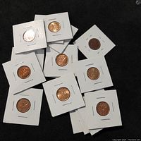 Top-down view of eleven Canadian 1 cent coins individually mounted in white cardboard holders with staples, arranged on a dark surface.