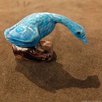 Close-up angled top view of blue porcelain bird figurine on brown base, showing feather texture and glossy glaze.