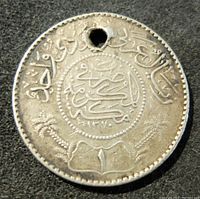 Front side of old Saudi Arabia 1 Riyal silver coin with hole drilled near the top, showing Arabic inscriptions, palm fronds, and denomination '1'.