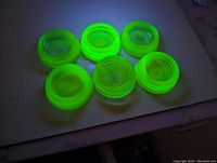Top view of six signed Hazel Atlas Vaseline glass furniture coasters glowing under UV light showing bright fluorescent green color and round shape.