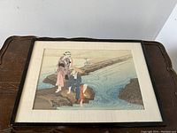Framed woodblock print depicting two women by a stream on a wooden bridge, photographed from front on a wooden table.