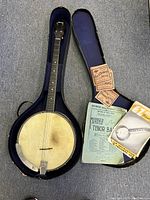 Full view of antique tenor banjo in original open hard case with extra strings packages, tuning fork, and song books.