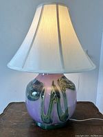 Full view of the table lamp lit, showing the white fabric shade and purple glass base with green and black abstract swirl patterns.