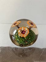 Front view of the clear glass paperweight with orange Millefiori mushrooms inside on a green base.