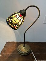 Table lamp with curved metal stem and textured circular metal base, featuring a stained glass lampshade with geometric patterns and colorful jewel accents, lit and off to show detail.
