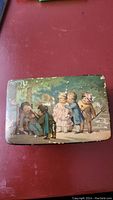 Rectangular metal musical box in painted tin case with a vintage illustration of children playing outdoors, showing wear and paint loss.