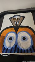 Oil on paper painting showing a stylized abstract face or mask with prominent eyes and geometric shapes.