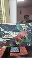 Full view of one abstract painting on board showing a human figure entwined with a green snake and natural elements.