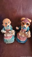 Two vintage bear toys dressed in fabric clothing with small plush bears, placed on brown leather surface.