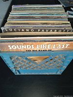 Photo showing a blue milk crate filled with stacked vinyl records. Album covers visible include Luciano Pavarotti and Sounds Like Last by The Big Band
