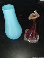 Photo showing the two art glass vases side by side on a black surface, one light blue and teardrop shaped, the other cranberry with clear ruffled top and base.