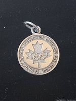 Front view of the round sterling silver pendant charm showing the maple leaf with the Olympic rings and text around the border on a black background.