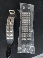 Photo shows two black leather punk style bracelets with silver-tone metal studs. One bracelet has large pyramid studs in two rows, the other has small square studs in multiple rows and is partially wrapped in plastic.