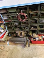 Wide angle view of heavy metal framed storage unit with small green drawers, antique ball vise, and red container with metal parts on floor.
