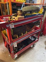 Red metal rolling tool cart with three shelves holding various tools and parts, showing dents and scratches.