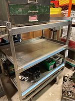 Metal cart showing three flat shelves with raised edges and caster wheels at the bottom