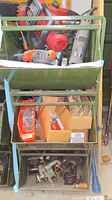 Green metal shelving unit with top shelf containing a Sears/Craftsman angle grinder, BD sander, and other tools; middle shelves with boxed small parts and packaging; bottom shelf with vintage mechanical tools in green metal casings and a long handled tool.