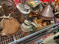 Top view showing a variety of oil cans and funnels arranged on metal shelving, highlighting materials such as copper and steel with weathered surfaces.
