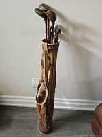 Full view of antique leather pipe golf bag standing upright with several hickory shaft clubs inside.