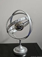 Front angled view of the metal armillary sphere globe on a black surface with grey background showing the spherical bands and reflective center globe.