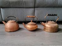 Three copper kettles displayed side by side on a gray floor, showing varying shapes, sizes, and handles.