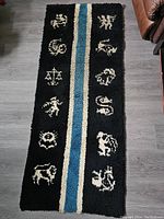 Full view of the rectangular black carpet or wall hanging showing twelve white zodiac symbols arranged around a central blue and cream stripe.