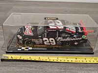 Front/side angle view of the Kevin Harvick #29 Budweiser diecast car in clear plastic display case on a wood surface next to measuring tape showing about 8.5 inches length.