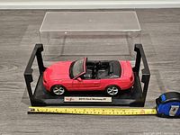 Front side angle photo showing red 2010 Ford Mustang GT convertible diecast model with black interior on display base with clear cover in background and measuring tape for scale.