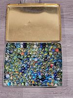 Open metal tin filled about half full with assorted glass marbles in various colors and patterns.