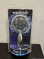 Front view of Waterpik Power Spray+ handheld shower head in sealed retail packaging showing chrome finish and spray nozzle arrangement.
