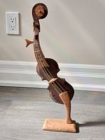Wooden string instrument carving figurine standing on a wooden base, photographed against a wall and floor background with electrical outlet visible.
