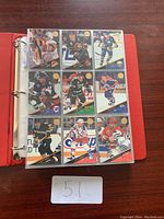 Photo showing a red binder open to a page of hockey cards in clear plastic sleeves; nine cards visible, featuring various players in NHL jerseys predominantly from 1990s-2000s era.