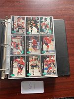 Page of hockey cards in a plastic sleeve showing multiple players in various team uniforms in a black three-ring binder on wooden surface.
