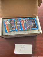 Photo showing an open box filled with a large number of colorful baseball trading cards, with several cards displayed on top featuring players in batting and fielding poses.