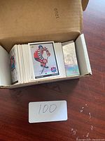 Photo showing open cardboard box filled with hockey cards, with some cards visible on top including one featuring a hockey player in team gear and a holographic card to the right.