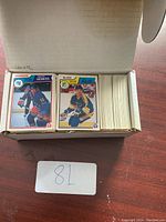 Open cardboard box showing stacks of hockey cards, with Anders Hedberg Rangers card and Blues Muller card visible on top.