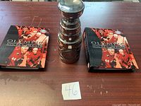 View of the Stanley Cup replica trophy and two Olympic Hockey Heroes booklets on a wooden surface with lot number card.
