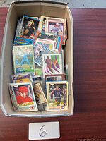 Overhead view of cardboard box filled with assorted sports trading cards: hockey, baseball, basketball; visible O-Pee-Chee Calgary Flames and Winnipeg Jets cards, a Panini Illusions basketball card, assorted other cards.