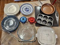 View of all items including Pyrex glass dishes, CorningWare covered dish, muffin tray, dinner plates, small covered dishes, fry pan, and baking sheet on hardwood floor.