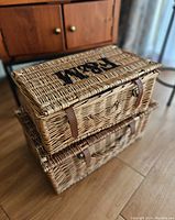 Two natural wicker baskets stacked, top basket smaller with 'HOME' text on lid, leather-like strap closures, woven handles visible