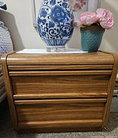 Photo of one matching nightstand in light wood-like veneer with 2 drawers and rounded edges, topped with a blue and white vase and a green pot with pink flowers.