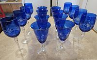 Overall top view of 8 blue wine glasses and 8 goblets arranged together on counter showing shape and detailing.