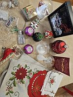 Photo of assorted Christmas decorations including snow globes with white tree and deer, small garlands with poinsettia, various baubles, placemats, and a red ornament box.
