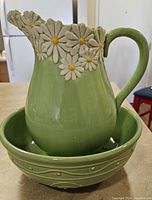 Green ceramic jug with a spout and handle, decorated with white daisies and yellow centers around the rim, placed inside a matching green bowl with textured details.
