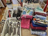 Wide shot showing full lot with cutlery in white plastic organizer, black handled utensils, and stacked dish towels with various patterns and colors.