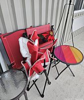 Photo showing 2 red folding chairs, one with floral cushion, 1 red and white Canada folding camp chair, clear glass top side table and multi-colored rainbow side table.